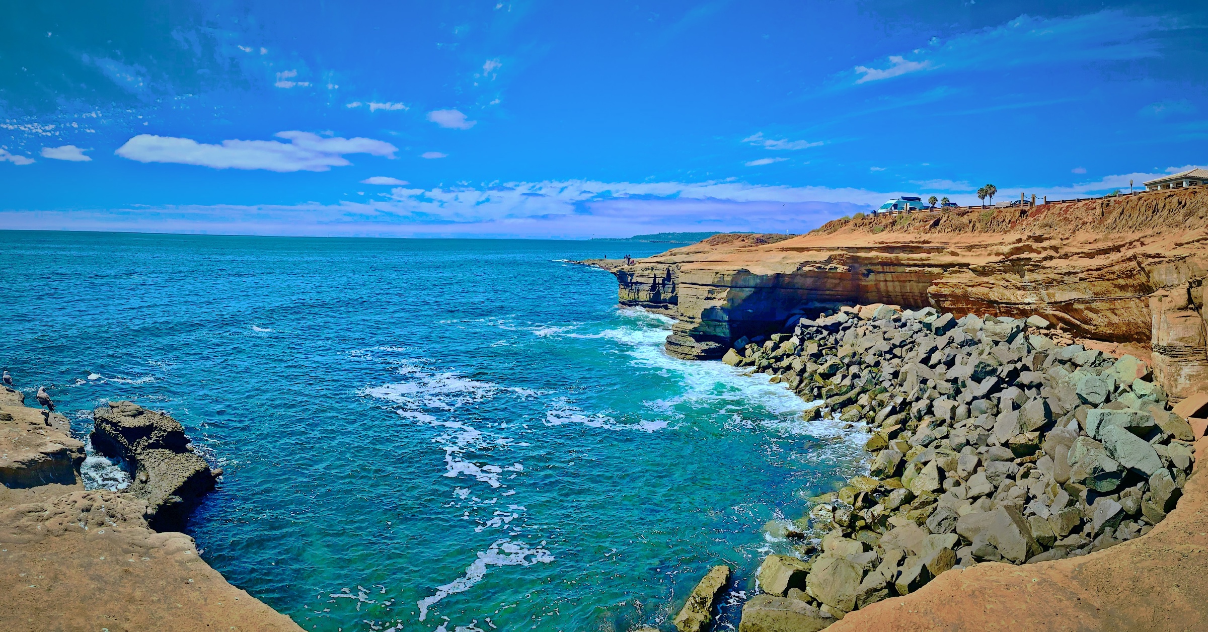 A wide panoramic view of turquoise ocean water meeting steep sandstone cliffs and stacked grey boulders under a bright sky.