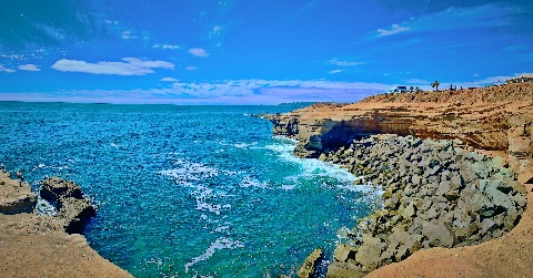 A wide panoramic view of turquoise ocean water meeting steep sandstone cliffs and stacked grey boulders under a bright sky.