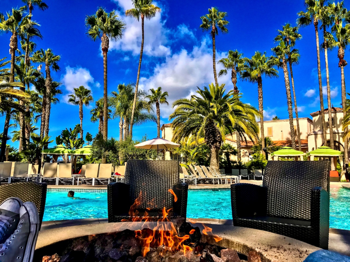 A fire pit with active flames sits poolside near dark woven chairs, surrounded by tall palm trees under a bright blue sky with white clouds.