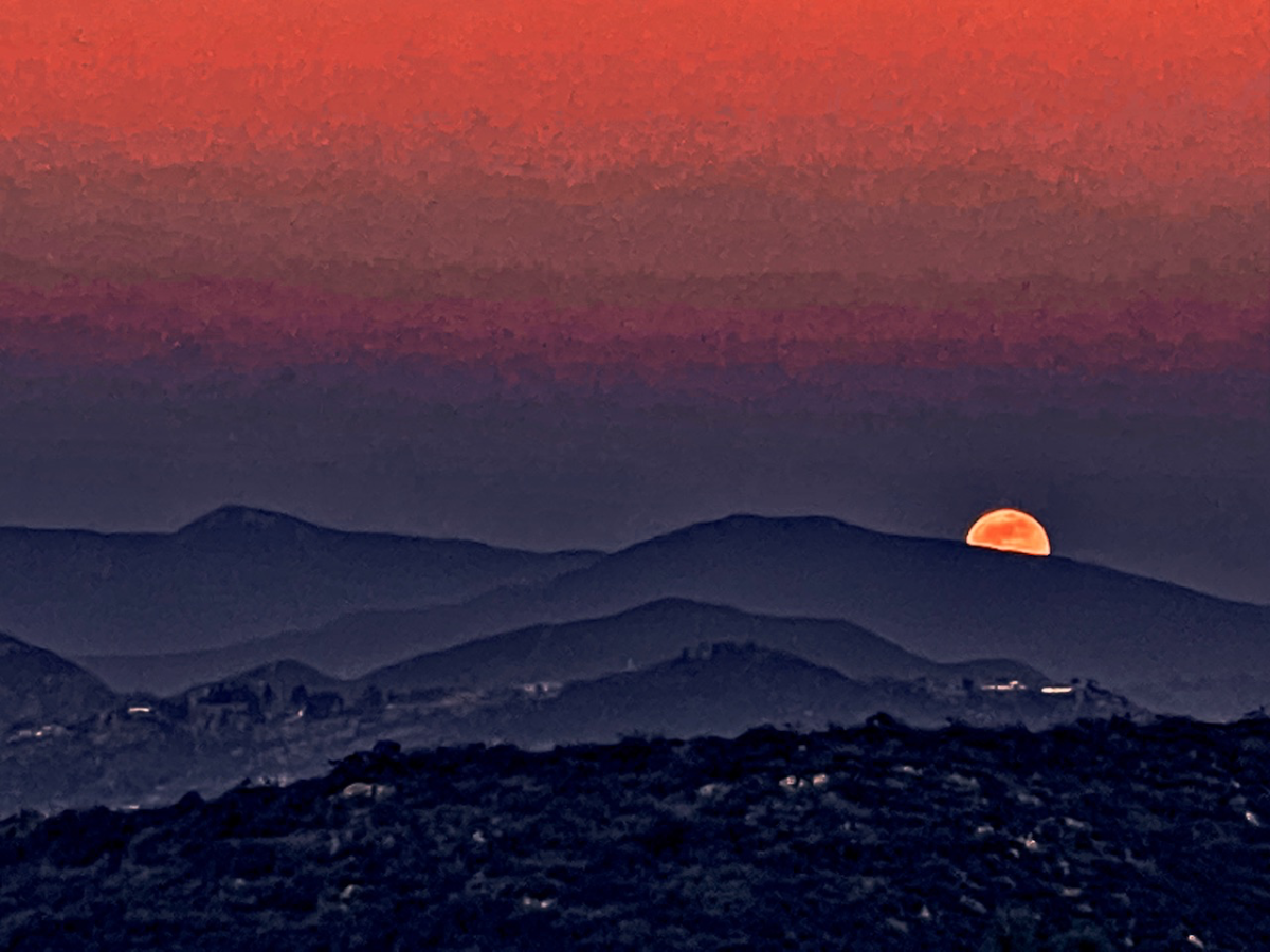 A glowing orange moon dips behind layered dark mountain ridges under a vivid red sky.
