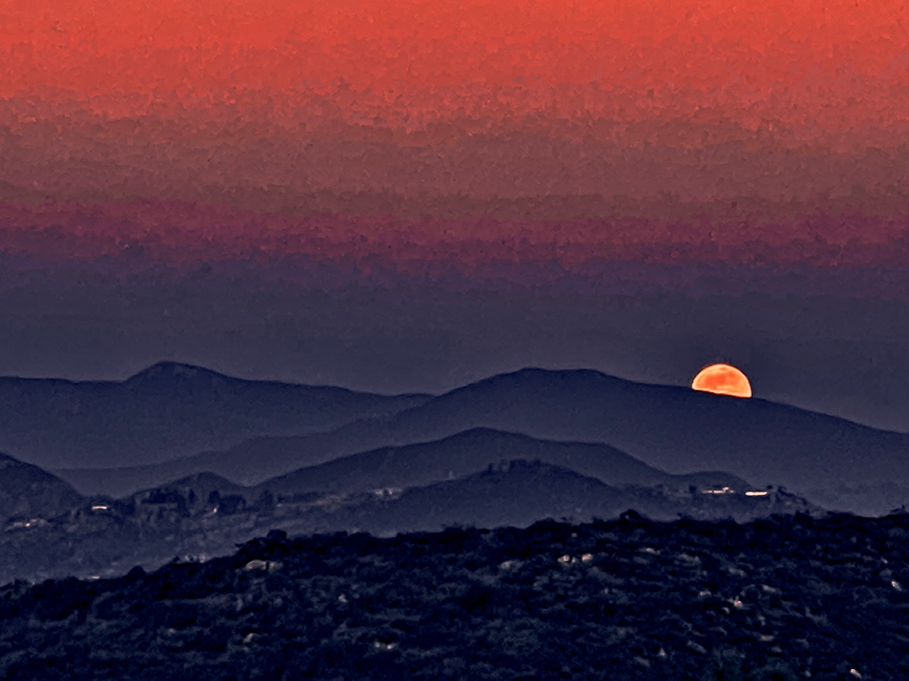 A glowing orange moon dips behind layered dark mountain ridges under a vivid red sky.