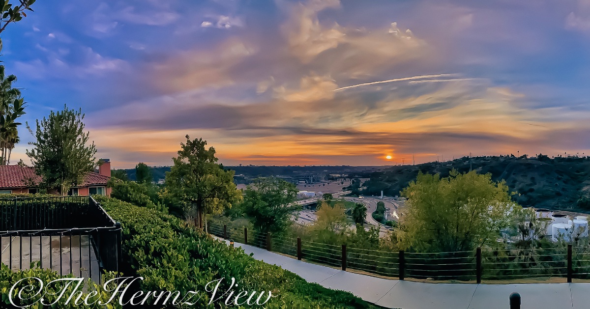 A paved walking path along a terraced hillside overlooks a valley and highway network during a vibrant orange and purple sunset.