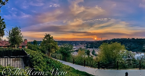A paved walking path along a terraced hillside overlooks a valley and highway network during a vibrant orange and purple sunset.
