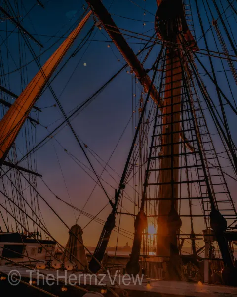 Intricate wooden ship masts, ropes, and ratlines silhouetted against a gradient sunset sky fading from dark blue to warm orange.