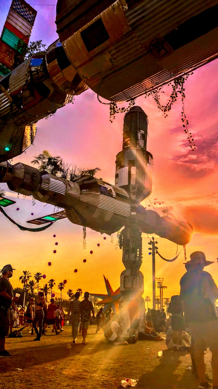 Low-angle view of large metal art installations shaped like rockets against a vivid pink and orange sunset with silhouetted people walking below.