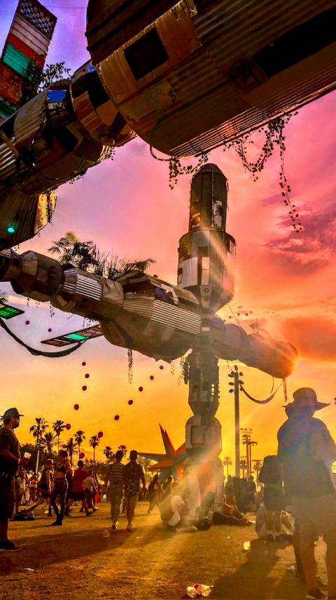Low-angle view of large metal art installations shaped like rockets against a vivid pink and orange sunset with silhouetted people walking below.