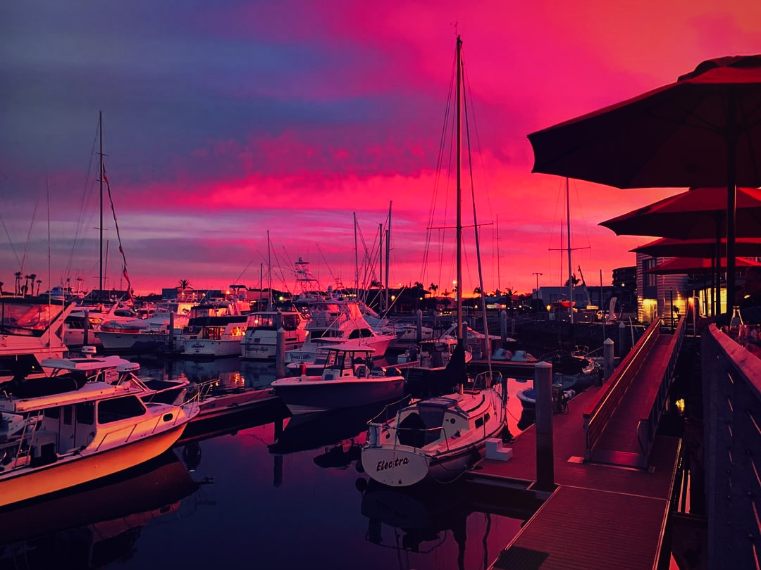 Docked boats and sailboats in a calm marina under a vivid magenta and purple sunset sky, with patio umbrellas visible on the right.
