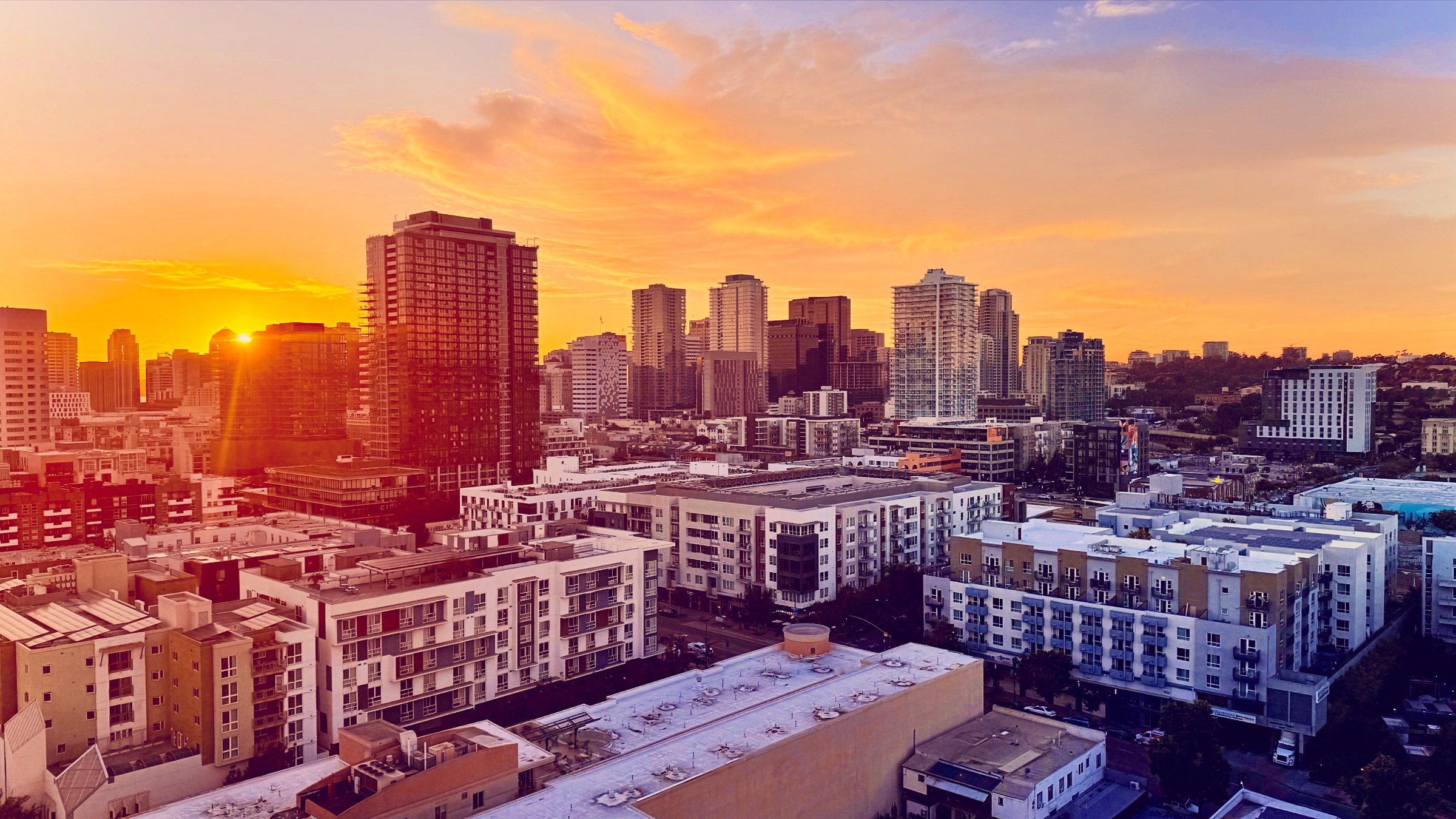 An elevated view of a city skyline at sunset, with the sun bursting beside high-rise buildings and casting a bright golden orange light across the urban rooftops.