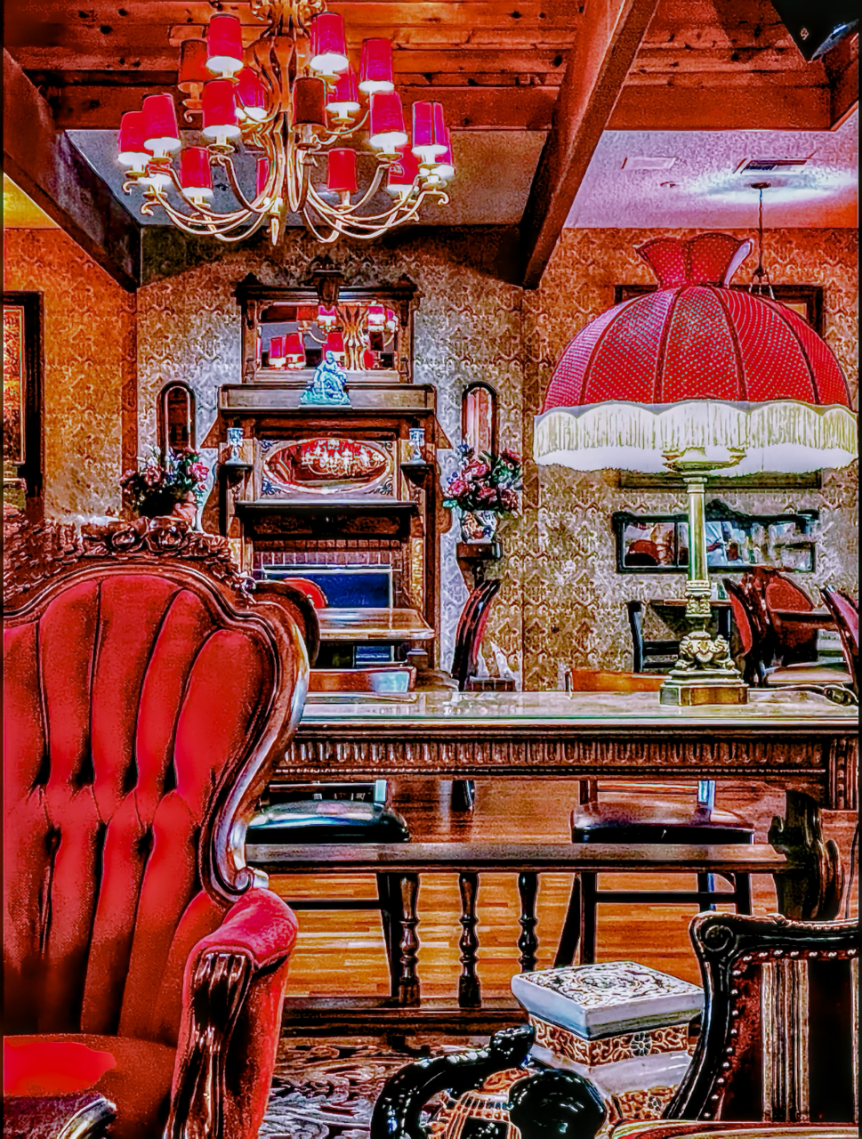 A richly decorated interior room featuring red lampshades, a central chandelier, patterned wallpaper, a red velvet armchair, and carved dark wood furniture.