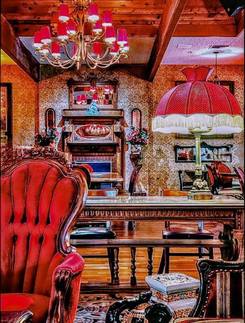 A richly decorated interior room featuring red lampshades, a central chandelier, patterned wallpaper, a red velvet armchair, and carved dark wood furniture.