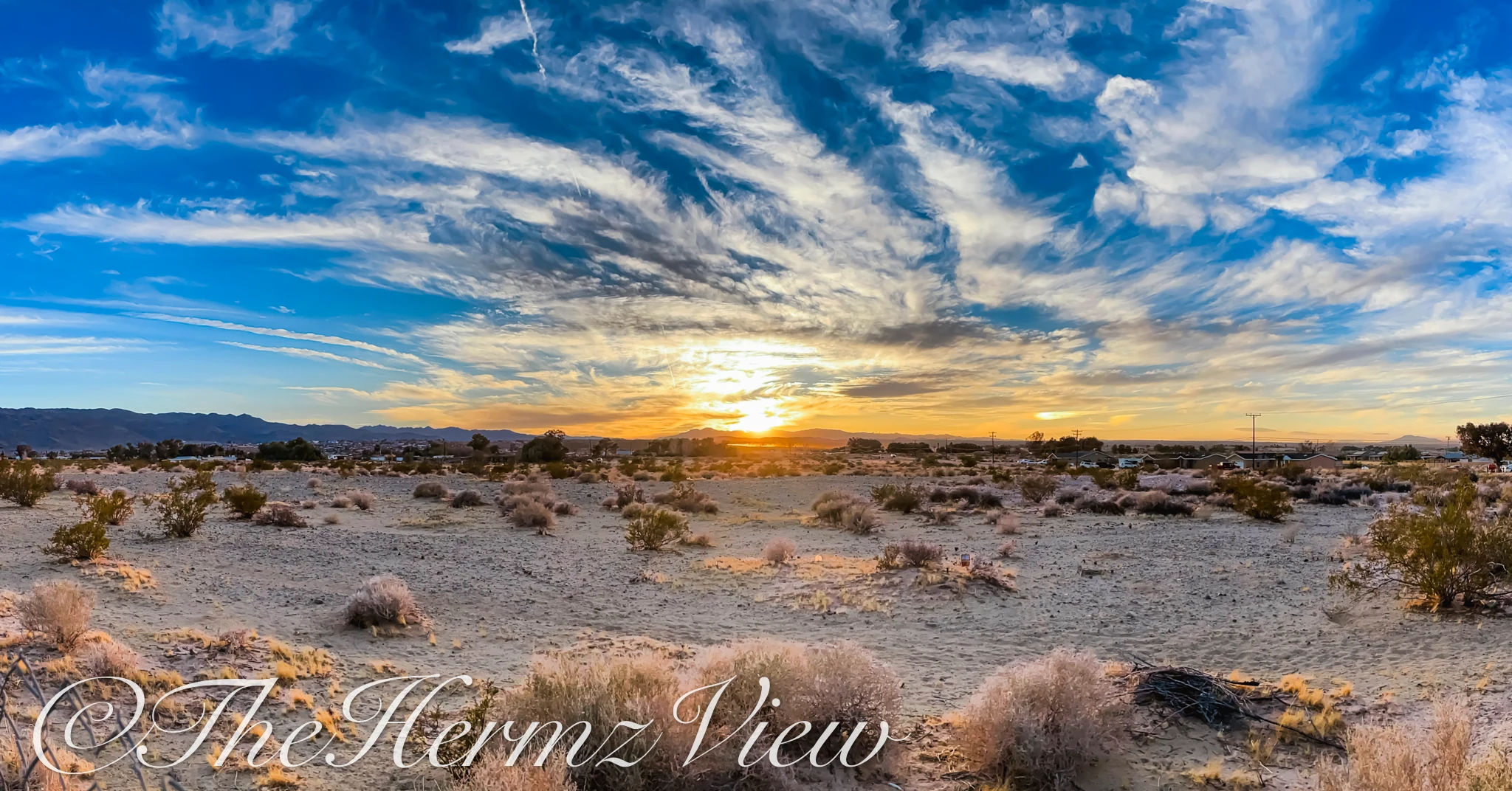 Wide panoramic view of sparse desert shrubs across sandy terrain under a blue sky with wispy clouds surrounding the setting sun on a mountain horizon.