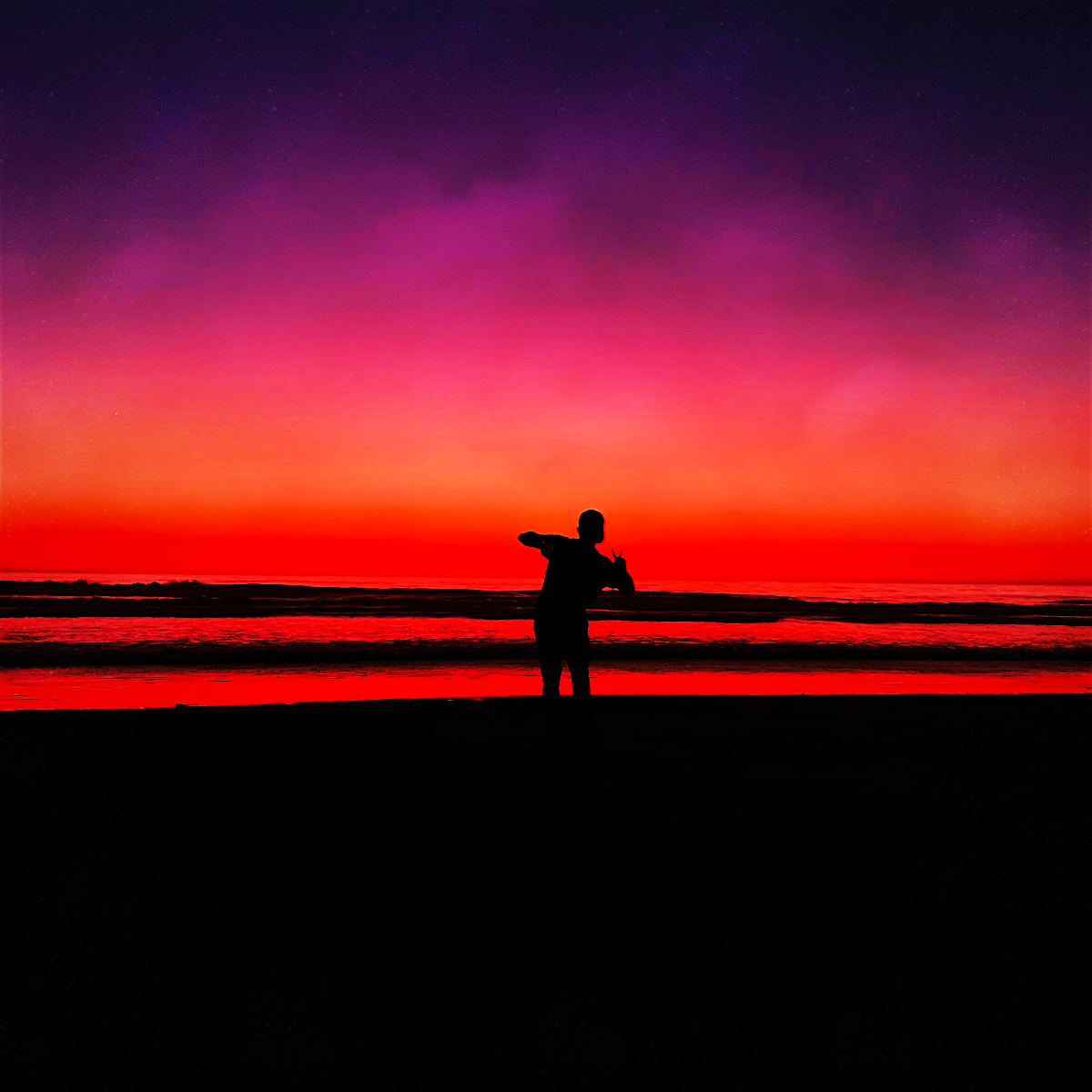Silhouette of a person standing on a sandy shore under a vivid purple, pink, and red sunset sky.
