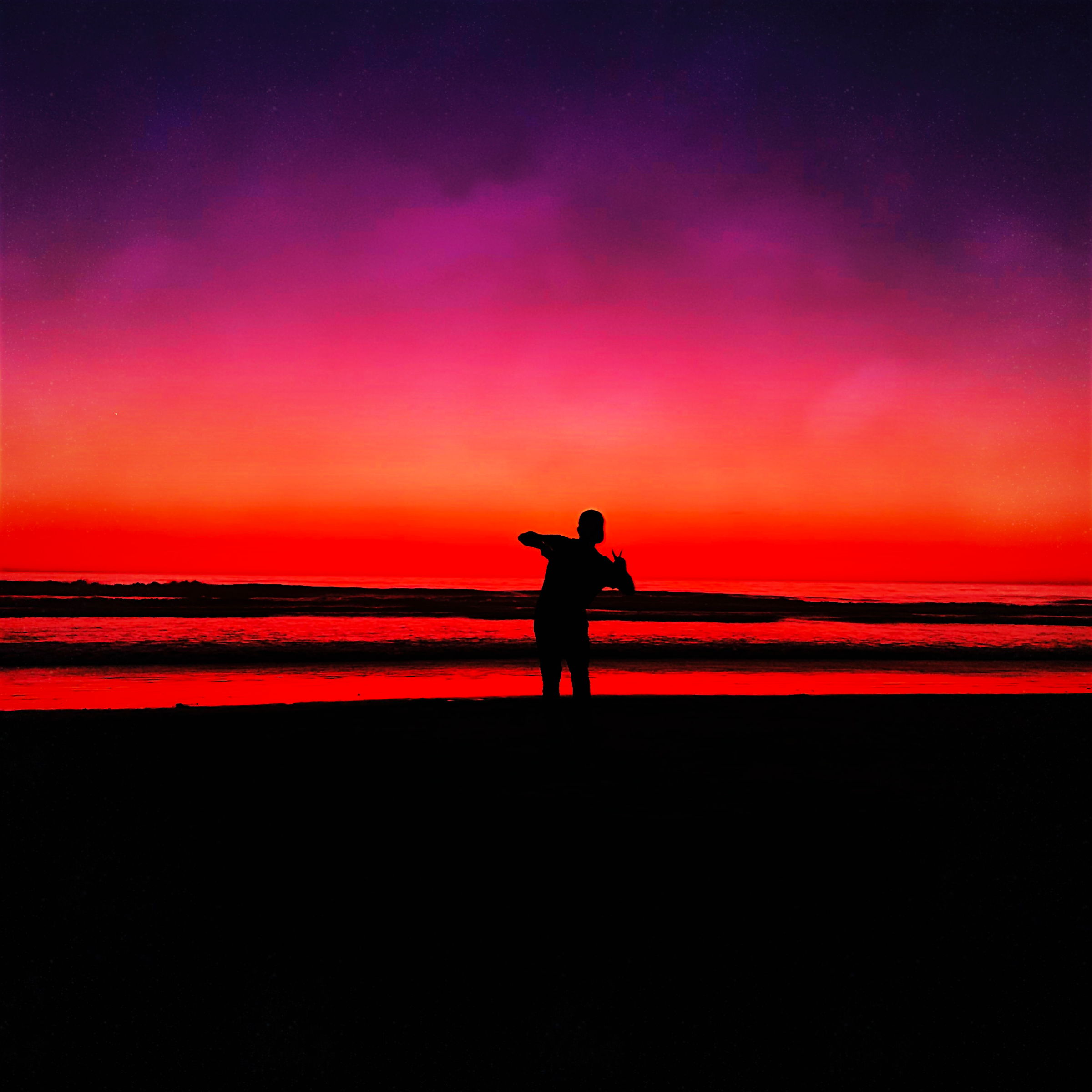 Silhouette of a person standing on a sandy shore under a vivid purple, pink, and red sunset sky.