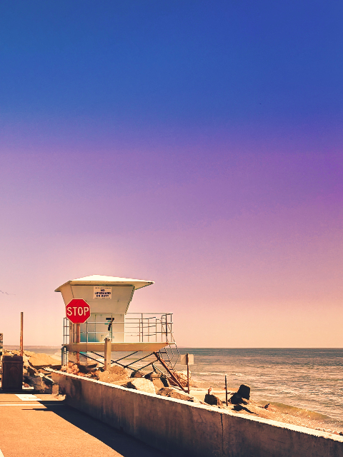 Lifeguard tower number 7 stands beside a red stop sign along a concrete seawall overlooking the ocean under a blue and pink gradient sky.