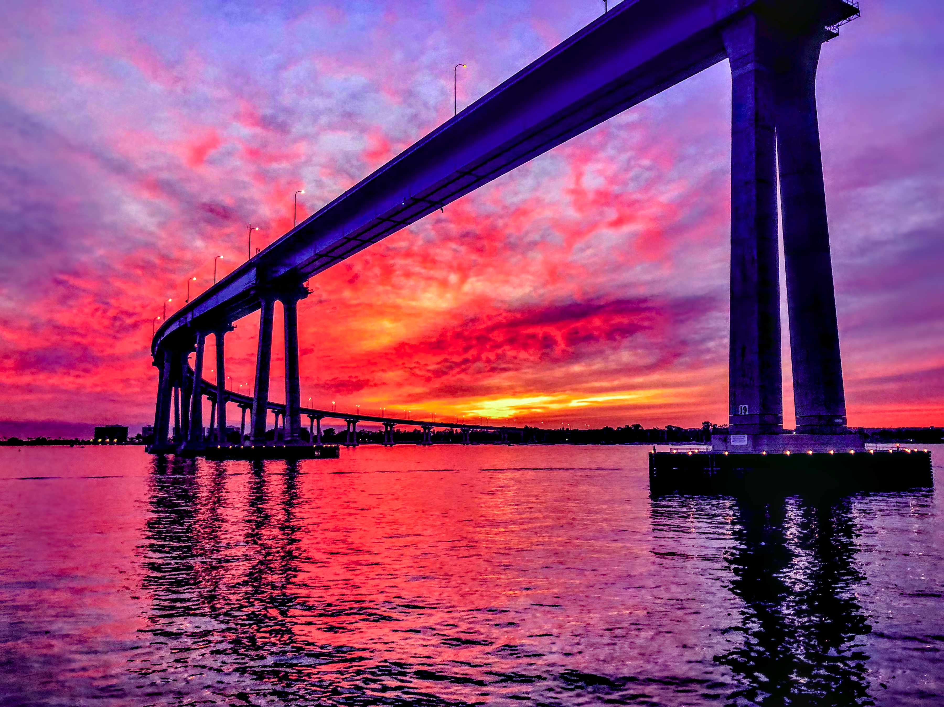 A long curved bridge supported by concrete piers extends over water against a sky filled with vivid magenta, purple, and orange twilight clouds.