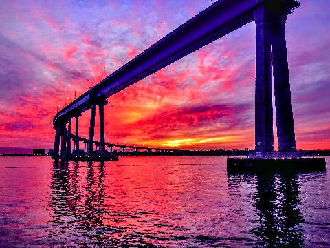 A long curved bridge supported by concrete piers extends over water against a sky filled with vivid magenta, purple, and orange twilight clouds.