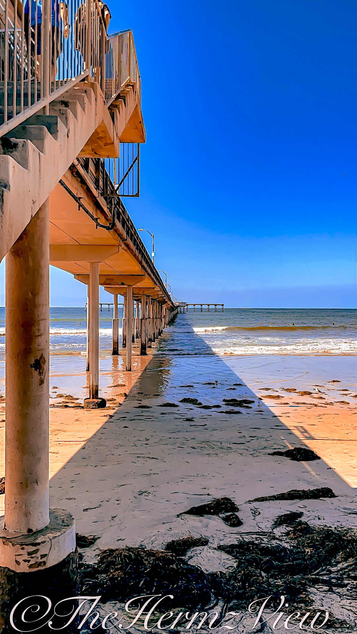 Low-angle perspective beneath a concrete ocean pier, casting a sharp geometric shadow across wet sand scattered with seaweed under a bright blue sky.