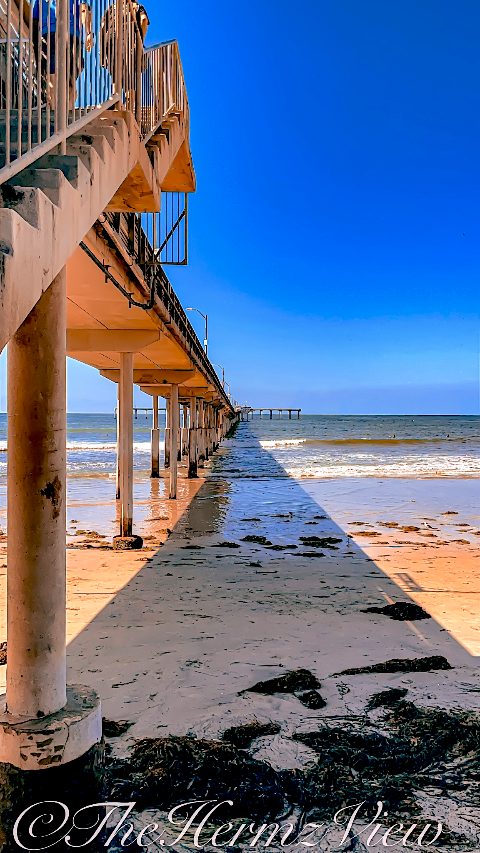 Low-angle perspective beneath a concrete ocean pier, casting a sharp geometric shadow across wet sand scattered with seaweed under a bright blue sky.