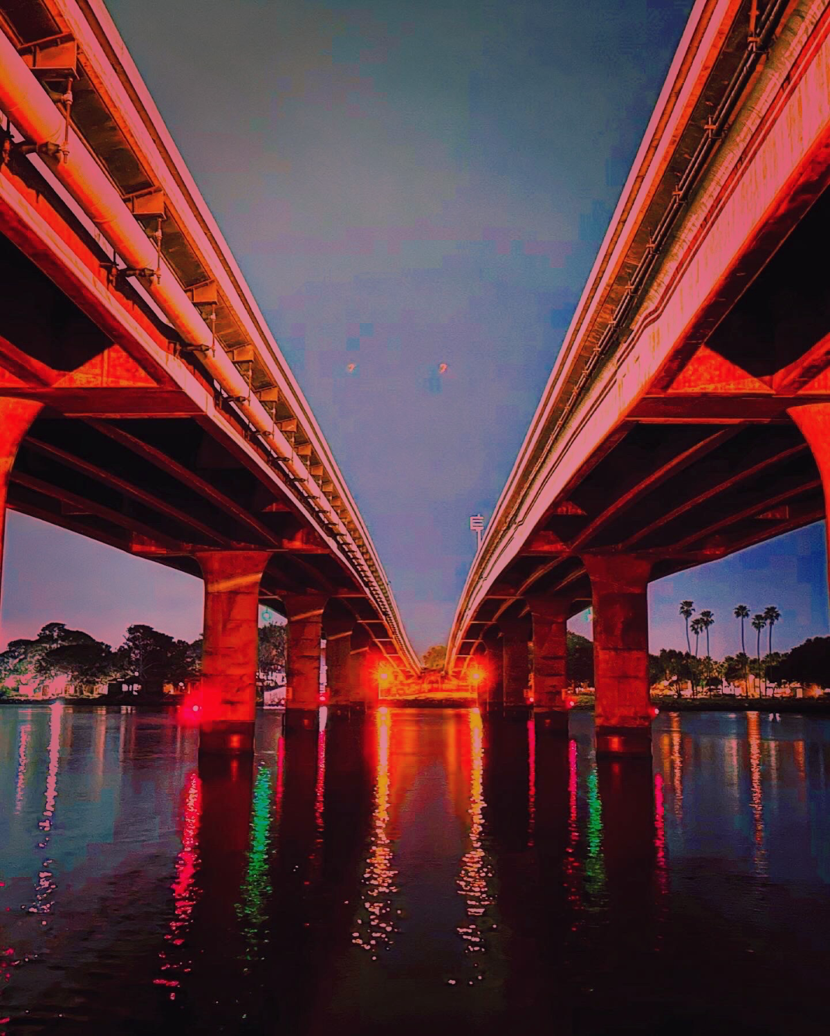 Low-angle perspective looking down a body of water situated between two parallel concrete bridge decks lit with a red glow at twilight, reflecting vibrant lights in the water.
