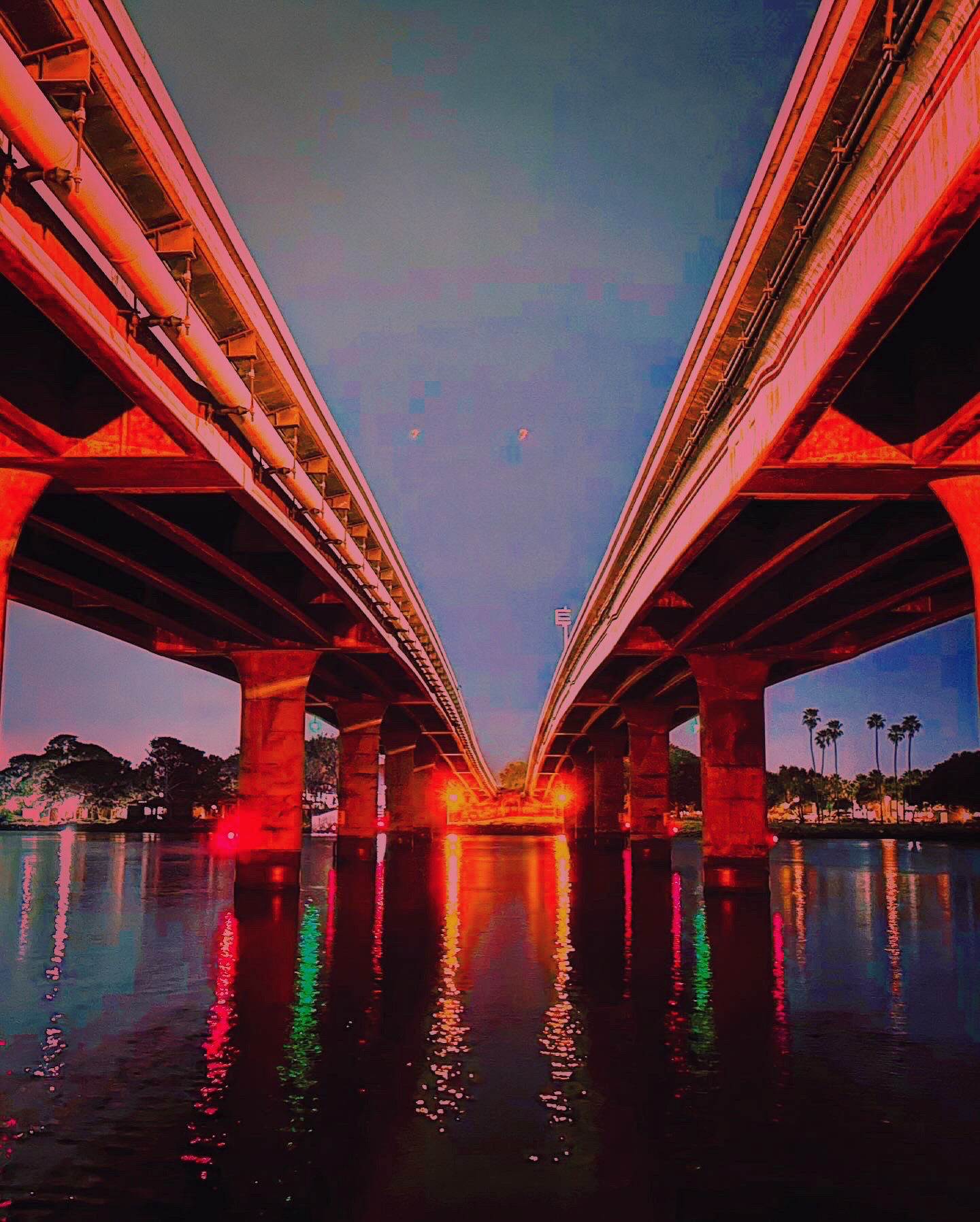 Low-angle perspective looking down a body of water situated between two parallel concrete bridge decks lit with a red glow at twilight, reflecting vibrant lights in the water.