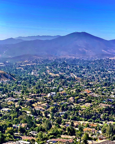 High-angle view looking down over a lush, tree-filled suburban valley toward distant mountain ridges under a bright blue sky.