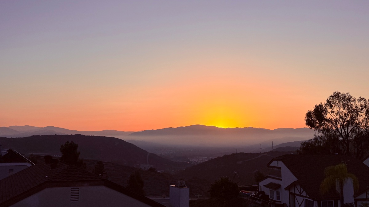 Silhouetted mountain ridges and suburban rooftops under an orange and soft purple gradient sky with bright light glowing over distant hills.