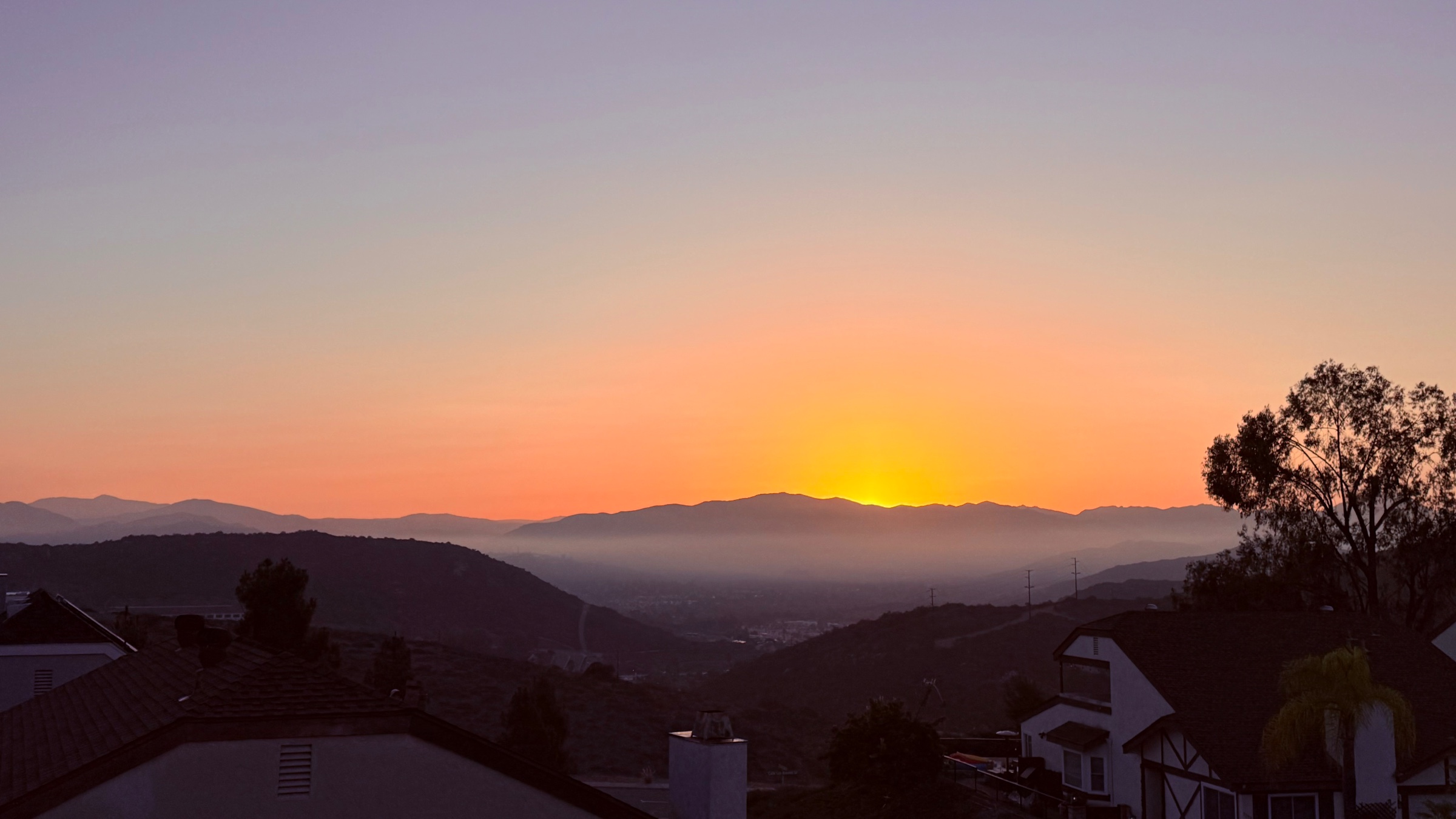 Silhouetted mountain ridges and suburban rooftops under an orange and soft purple gradient sky with bright light glowing over distant hills.